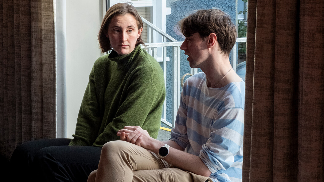 Two people sit by a window having a serious conversation. One person wears a green sweater and looks at the other, who is dressed in a light blue and white striped shirt and beige pants, with hands resting on their lap.
