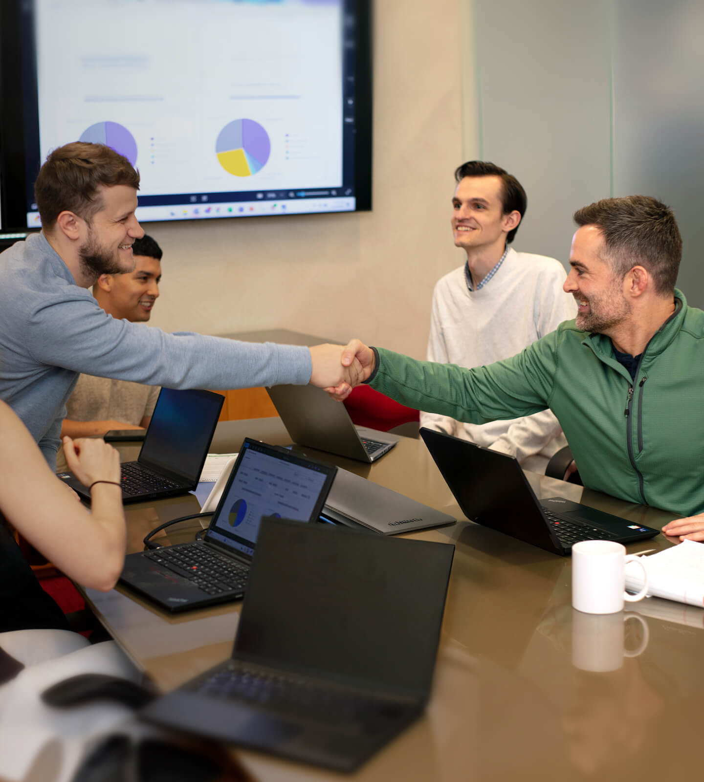 Four people in a meeting room sit around a table with laptops, while two men in the foreground smile and shake hands. A presentation with charts is displayed on a screen in the background.