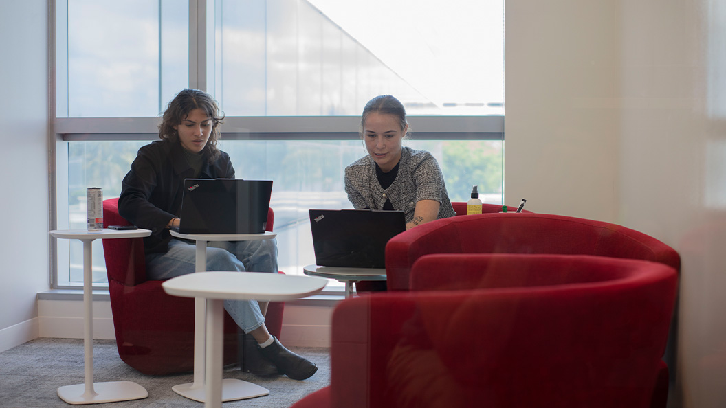 Two people sitting in red chairs at small round tables, working on laptops near a large window in a modern office space. Natural light fills the room.