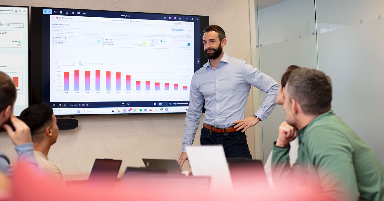 A man stands and presents data visualizations on a large screen to a group of people seated around a table with laptops in a modern office meeting room.